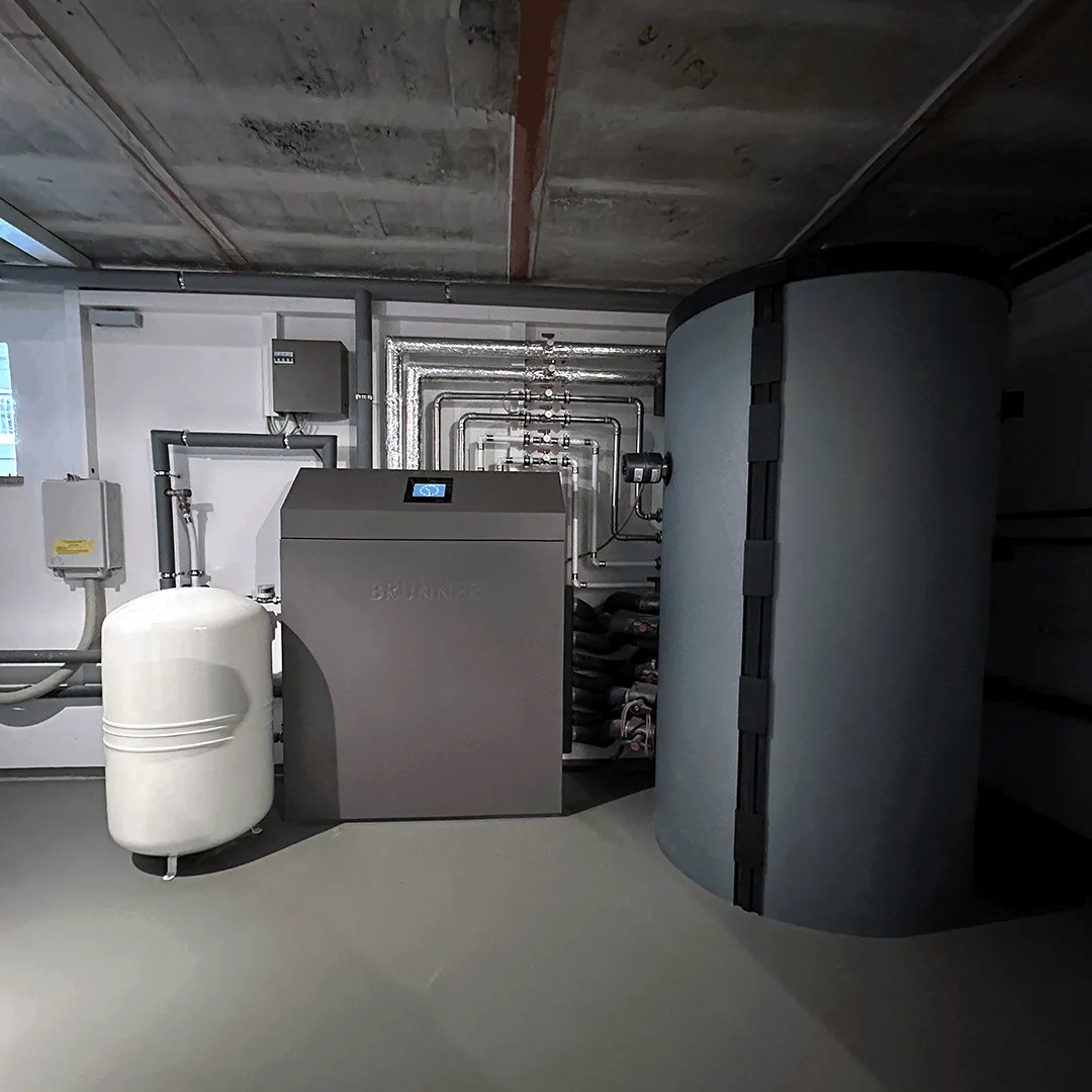 Das Bild zeigt den Heizraum im Keller mit einer modernen Heizungsanlage.  In der Mitte steht die kompakte, quaderförmige Heizzentrale in dunklem Grau mit einer kleinen digitalen Anzeige auf der Oberseite; der Schriftzug „BRUNNER“ ist dezent auf der Front erkennbar. Rechts daneben befindet sich ein großer, zylindrischer Pufferspeicher in dunkler Farbe, der deutlich höher ist als die Heizzentrale. Links im Bild steht ein weißer, runder Druckausgleichsbehälter auf kurzen Füßen.