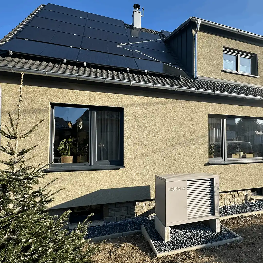 Das Bild zeigt die Außenansicht des Einfamilienhauses von Martin aus Zwickau. Das Haus hat eine helle, beige Fassade und ein schräges Dach, das fast vollständig mit dunklen Photovoltaikmodulen bedeckt ist. Auf dem Dach ist außerdem ein Schornstein zu sehen. Im Vordergrund, rechts neben dem Haus, steht auf einem kleinen Fundament die kompakte Wärmepumpe von Brunner.